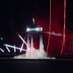 Witte Tesla Roadster zweeft met brandende koplampen boven de grond op vier rookpluimen van stuwraketten, tussen schijnwerpers en rode lichtlijnen tijdens de nachtelijke presentatie.
