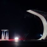 Tesla Roadster zweeft op vier stuwstralen boven de baan, naast een enorme gebogen schans van beton op een stalen stellage. Op de grond rijdt een cameravoertuig.
