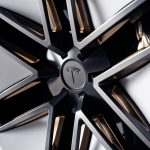 Close-up van de stervormige velg van de Tesla Roadster met Tesla-logo op de naafdop en koperkleurige accenten tussen de spaken.