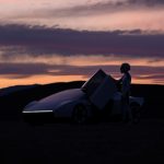 Coureur met helm opent de omhoog scharnierende deur van de Tesla Roadster bij zonsondergang, met heuvels en roze wolken op de achtergrond.